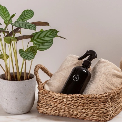Cesta de palha com toalhas e frasco spray preto ao lado de vaso com planta verde
