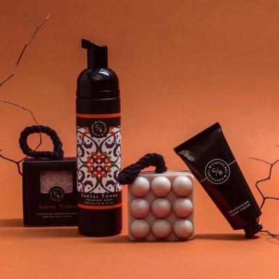 Produtos de cuidado corporal SANTAL TONKA em embalagens pretas com padrões laranja e branco, sobre fundo laranja com decoração de cordão preto e galhos secos.