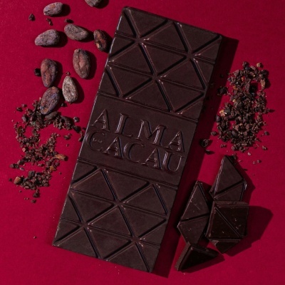 Tablete de chocolate negro ALMA CACAU com grãos de cacau ao lado em fundo vermelho