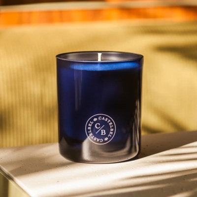 Vela perfumada em copo de vidro azul escuro com logotipo branco sobre mesa clara.