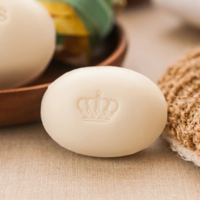 Sabonete branco oval com coroa gravada sobre superfície bege ao lado de esponja rústica e caixa de madeira.