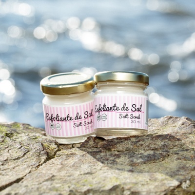 Dois frascos pequenos de esfoliante de sal com tampa dourada e rótulos rosa com branco sobre uma pedra junto ao mar