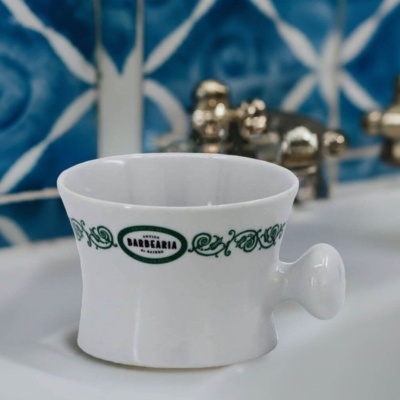 Copo branco em cerâmica com pega, padrão verde e texto Antiga Barbearia, sobre pia com azulejos azuis ao fundo.