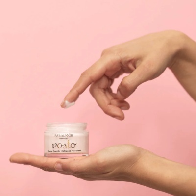 Frasco de creme facial BENAMÔR branco com texto rosio, segurado por mãos com fundo rosa