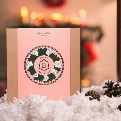 Caixa de papel castanho com rótulo rosa BENAMÔR ROSTO LISBOA 1925 em cenário natalício com neve artificial e pinhas.