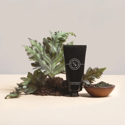 Tubo preto de creme para mãos ao lado de folhas verdes, terra e tigela de madeira com folhas secas