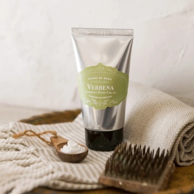 Creme de mãos Verbena em tubinho metálico prateado com etiqueta verde, sobre pano bege, com colher de madeira e escova de madeira