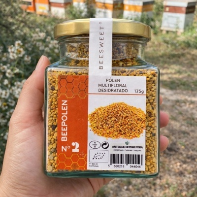 Frasco de vidro com pólen multifloral desidratado BEEPOLÉN 175g numa mão, com caixas de abelhas ao fundo