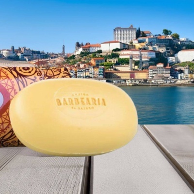 Sabonete amarelo oval com texto BARBEARIA em madeira clara com pano estampado e vista de cidade costeira