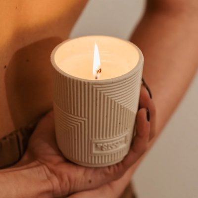 Vela acesa em recipiente de cerâmica bege com padrão geométrico, segura por mãos com unhas pintadas de preto.
