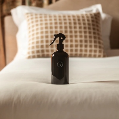 Frasco de spray preto numa cama com lençol branco e almofada castanha com padrão