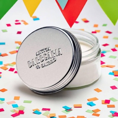 Pote de vidro com tampa metálica com gravação e creme branco rodeado por confetes coloridos e bandeirolas coloridas
