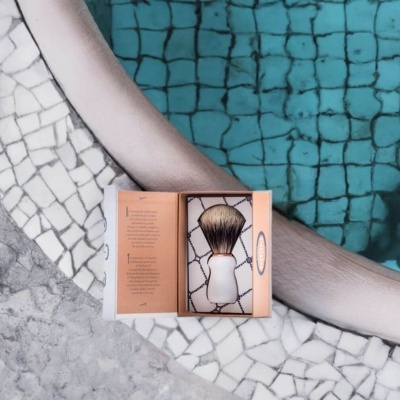 Pincel de barbear em embalagem aberta sobre mosaico branco junto a piscina