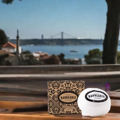 Sabonete Antiga Barbearia de Bairro com embalagem decorada castanha e preta sobre mesa de madeira com rio e ponte na paisagem ao fundo.