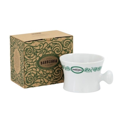 Tigela branca de cerâmica para barbear com pega e decoração verde junto à caixa de cartão castanha com padrão floral e texto Antiga Barbearia de Baixo