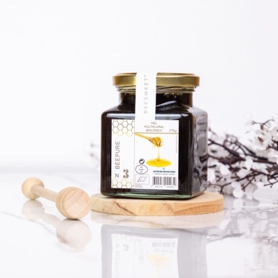 Frasco de mel multifloral biológico com tampa dourada em superfície branca com bastão de mel de madeira ao lado.