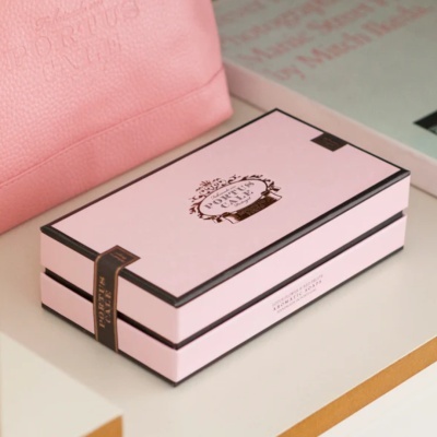 Caixa rosa retangular com bordas pretas e etiqueta preta, ao lado de saco rosa e livro cinza