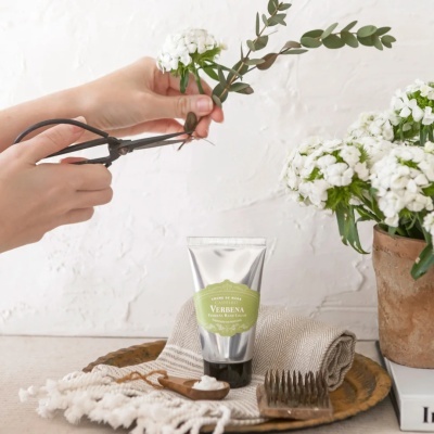 Flores brancas e verdes sendo cortadas com tesoura, creme de mãos Verbena, flores em vaso rústico e panos sobre mesa.