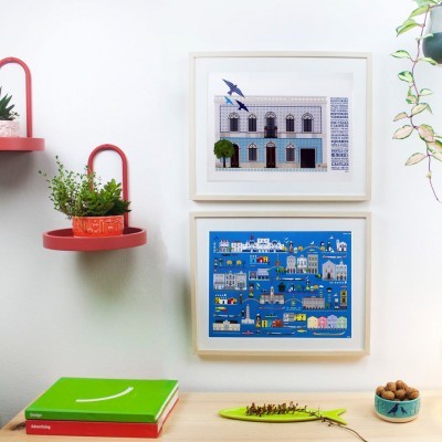 Duas ilustrações coloridas emolduradas numa parede branca junto a prateleiras vermelhas com plantas e itens decorativos numa mesa de madeira.