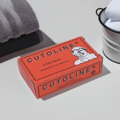 Caixa vermelha de produto Cutoline after shave, toalhas e balde branco em superfície branca
