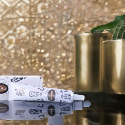 Tubo de creme branco com texto e design em preto, caixa branca e vasos dourados com plantas