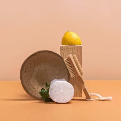 Escova de madeira, peça redonda branca com padrão de limão, prato castanho, limão amarelo e bloco de madeira em superfície laranja