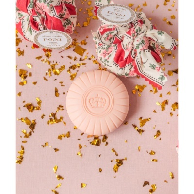 Barra de sabão rosa com relevo de coroa e embalagens em tecido floral sobre fundo rosa com confetes dourados