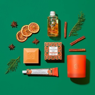 Produtos de beleza BENAMÔR LARANJINHA com óleo seco, sabonete, creme de mãos, vela e elementos naturais secados sobre fundo verde.