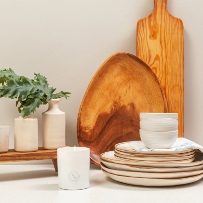 Louça branca com bordas castanhas empilhada, prato e tábua de madeira, vela branca acesa com CB, vasos de cerâmica bege com planta verde em fundo neutro.