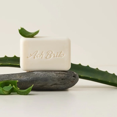 Sabonete branco com texto Ach Brito sobre pedra cinzenta e folhas de aloe vera verdes.