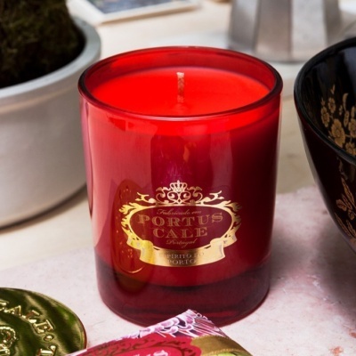 Vela vermelha em copo de vidro com etiqueta dourada numa superfície clara rodeada de objetos decorativos.