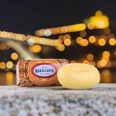 Sabonete amarelo e embalagem decorada Antiga Barbearia de Bairro sobre superfície cinzenta com fundo de luzes desfocado