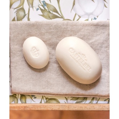 Duas barras de sabonete creme com escritas e símbolo, sobre pano bege e fundo floral.