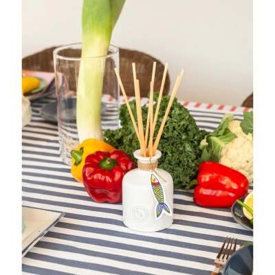 Difusor de aromas branco com pauzinhos de madeira e etiqueta em forma de peixe numa mesa com legumes frescos