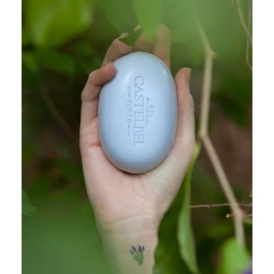 Sabonete oval azul claro CASTELBEL PORTO na mão com fundo verde e tatuagem pequena