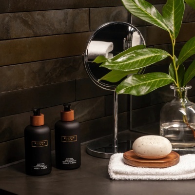 Duas embalagens de cosméticos pretas, sabonete branco sobre saboneteira de madeira, planta em jarro de vidro, e espelho redondo num balcão com azulejos escuros
