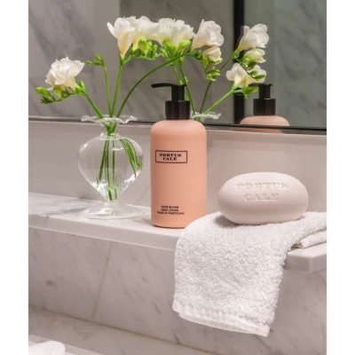 Frasco de loção corporal rosa, sabonete branco Portus Cale e vaso de flores brancas num balcão de casa de banho em mármore.