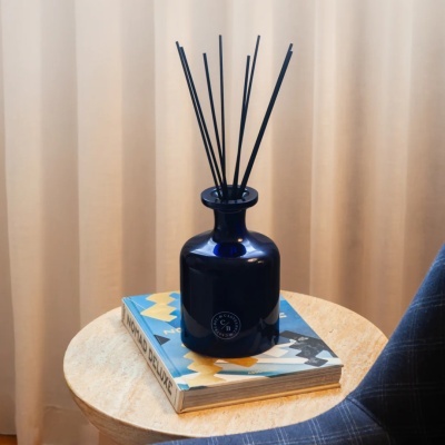 Difusor de ambiente azul escuro com varetas pretas sobre livro NOMAD DELUXE numa mesa redonda.