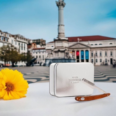 Navalha de barbear com cabo de madeira ao lado de caixa metálica prateada e flor amarela sobre mesa branca na rua