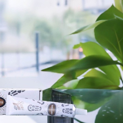 Tubo branco de produto cosmético com padrão de pegadas e rótulo Sardearia perto de caixa branca e folhas verdes