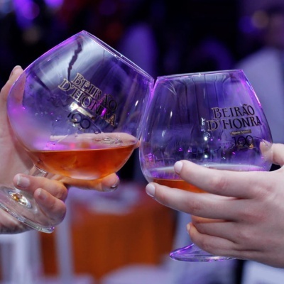 Duas mãos a brindar com copos de vidro com vinho licoroso e inscrições douradas BEIRÃO DHONRA.