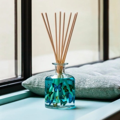 Difusor de ambiente em vidro com líquido azul e varetas de madeira junto a almofada e janela