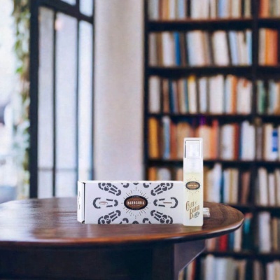 Frasco spray de óleo para barba e caixa de papel branco numa mesa de madeira com estantes de livros ao fundo.
