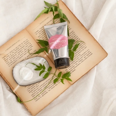 Creme de mãos Jasmim Branco em tubo prateado sobre livro aberto com folhas verdes e placa branca com creme.