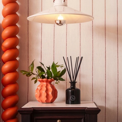 Mesa com vaso laranja e difusor preto, lâmpada pendurada e parede com riscas vermelhas