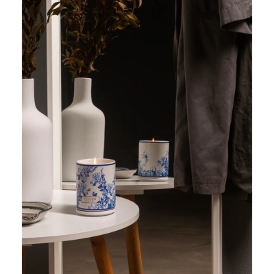 Vela decorativa branca com azul floral numa mesa branca ao lado de vaso com ramos secos