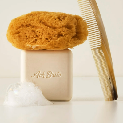 Esponja amarela, sabonete branco Ach. Brito e pente bege claro sobre fundo branco com espuma