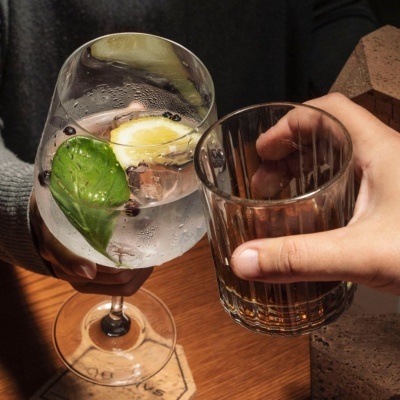 Mãos segurando dois copos de bebidas com gelo e decoração sobre mesa de madeira