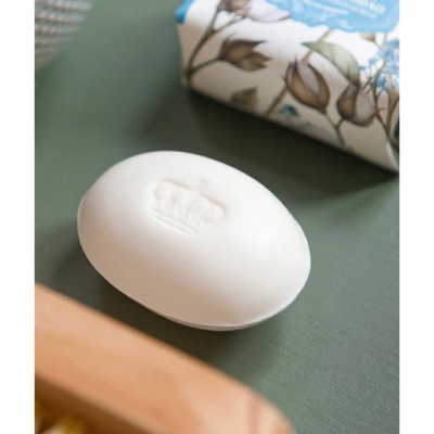 Sabonete branco oval com relevo de coroa sobre superfície verde com caixa floral ao fundo.