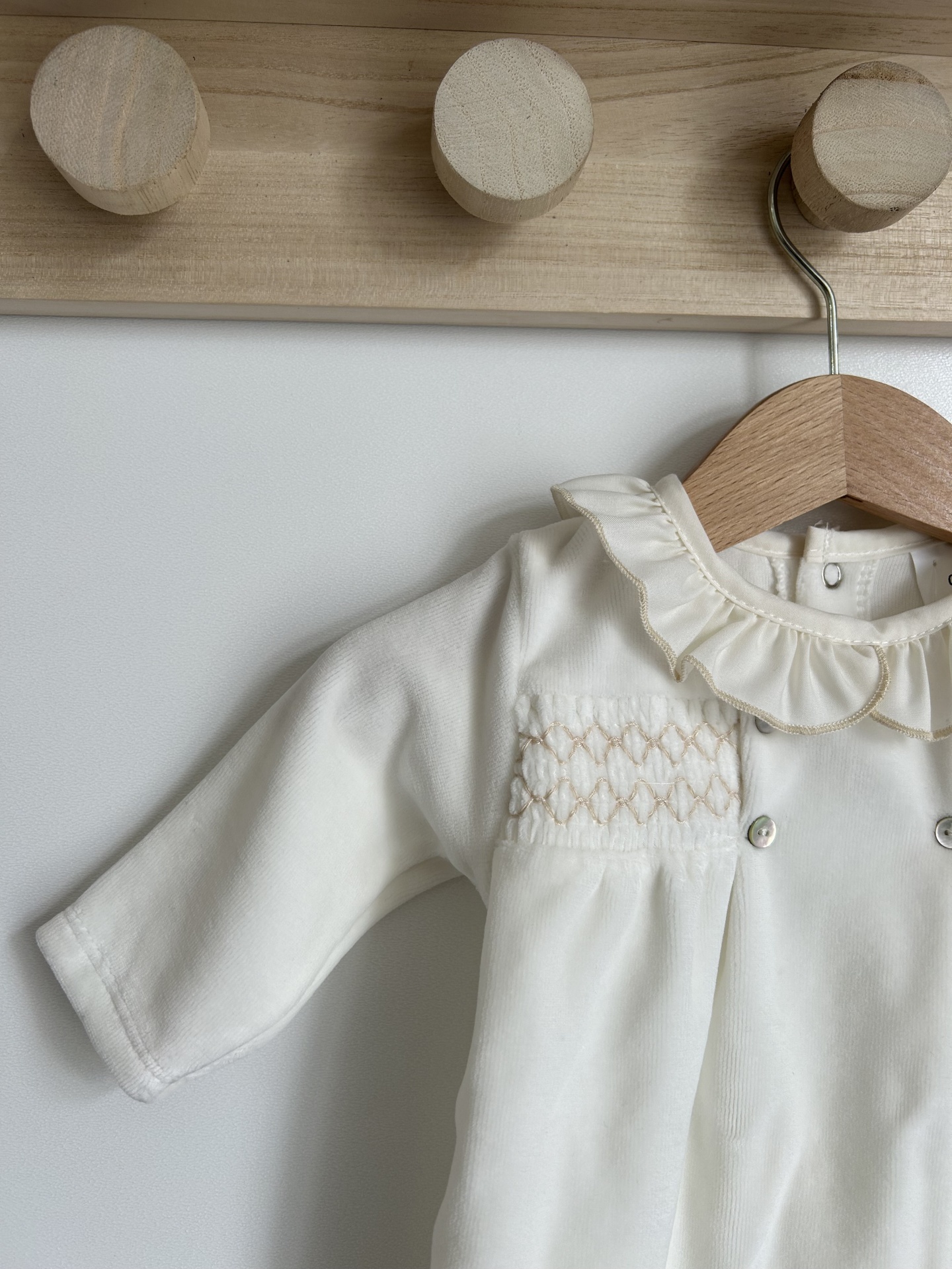 Macacão de bebé branco com gola folhos e bordados pendurado numa parede